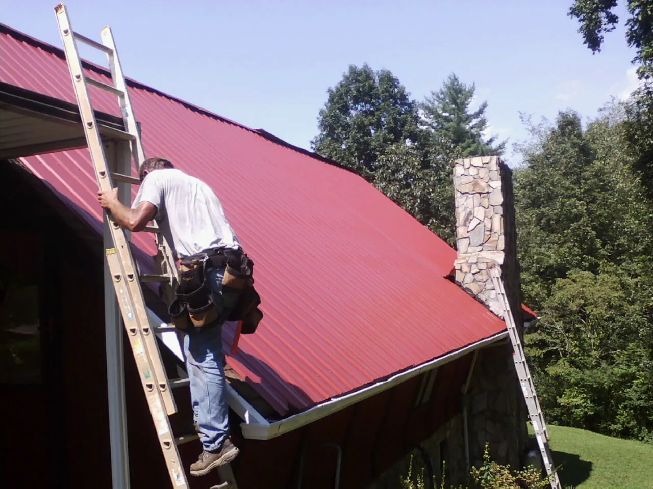 Roofer on a ladder beside a red metal roof and stone chimney — Soffit & Fascia Repair by Roofing Brothers in Levan